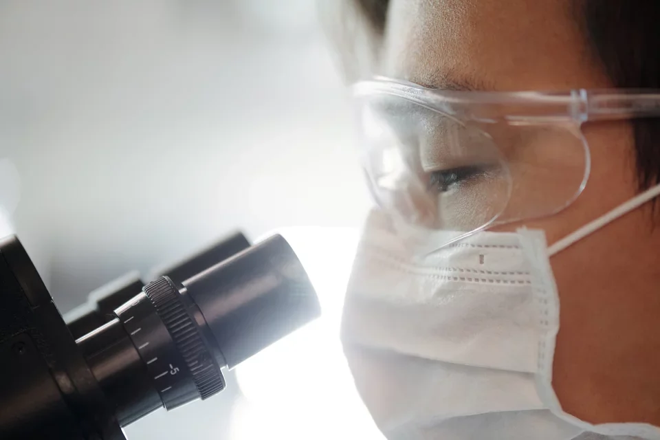 A researcher wearing protective glasses looks into a microscope.