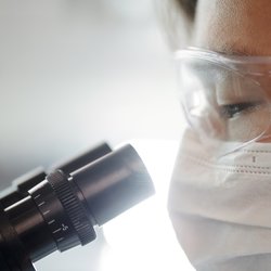 A researcher wearing protective glasses looks into a microscope.